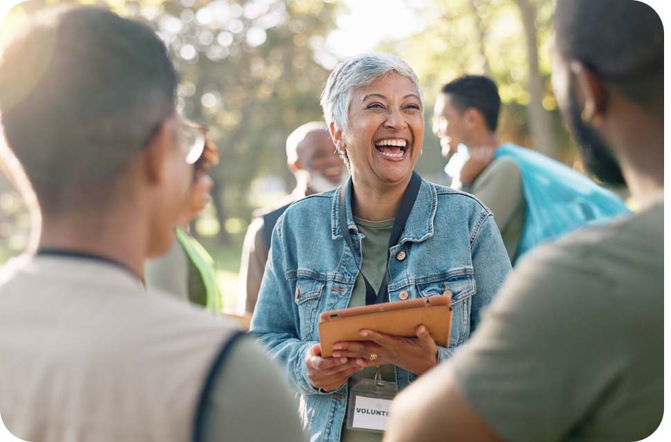 Older woman in denim jacket laughing, holding a notebook, chatting with students outdoors