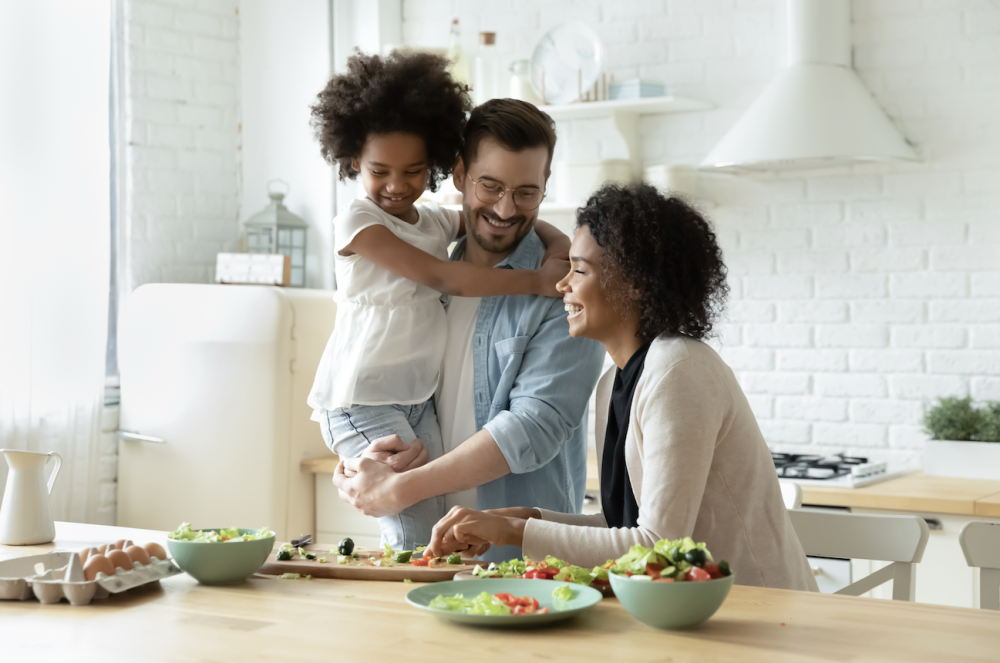 Smiling parents cooking, daughter on father's back in bright white kitchen