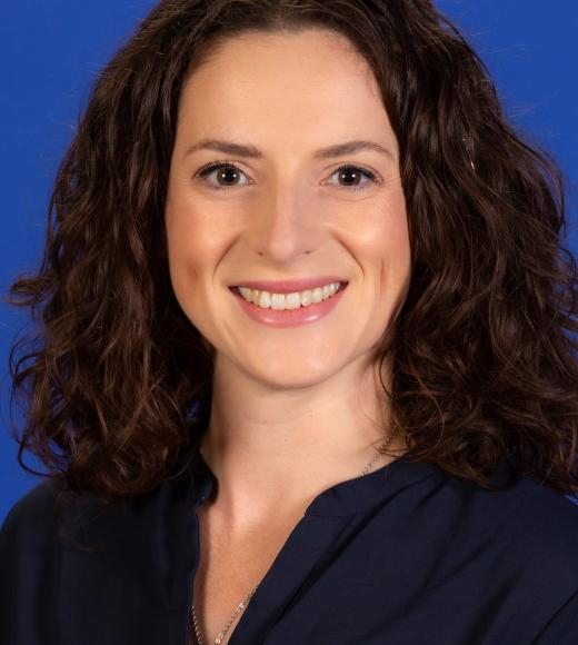 Professional headshot of smiling woman with curly brown hair against blue background