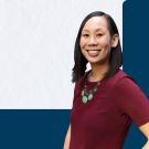 Smiling headshot of woman in maroon dress and green necklace on white and navy background
