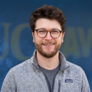 Headshot photograph of smiling young man with glasses and curly hair on blue textured backdrop