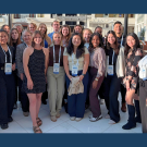 Diverse group of about 30 people smiling on a sunny outdoor terrace, wearing lanyards