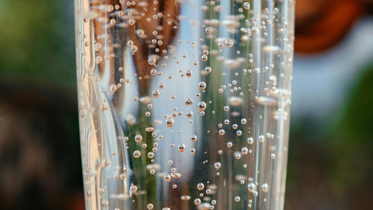 Clear glass of sparkling water on wooden table, bubbles rising, sunlit outdoor background