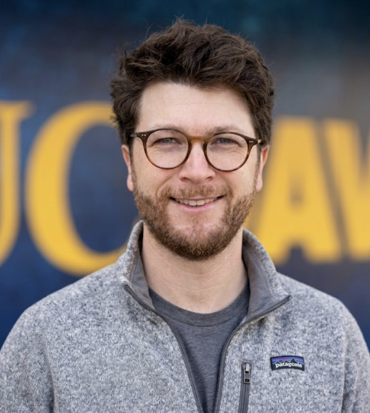 Headshot photo of a smiling bearded man with glasses against a blurred blue-yellow background
